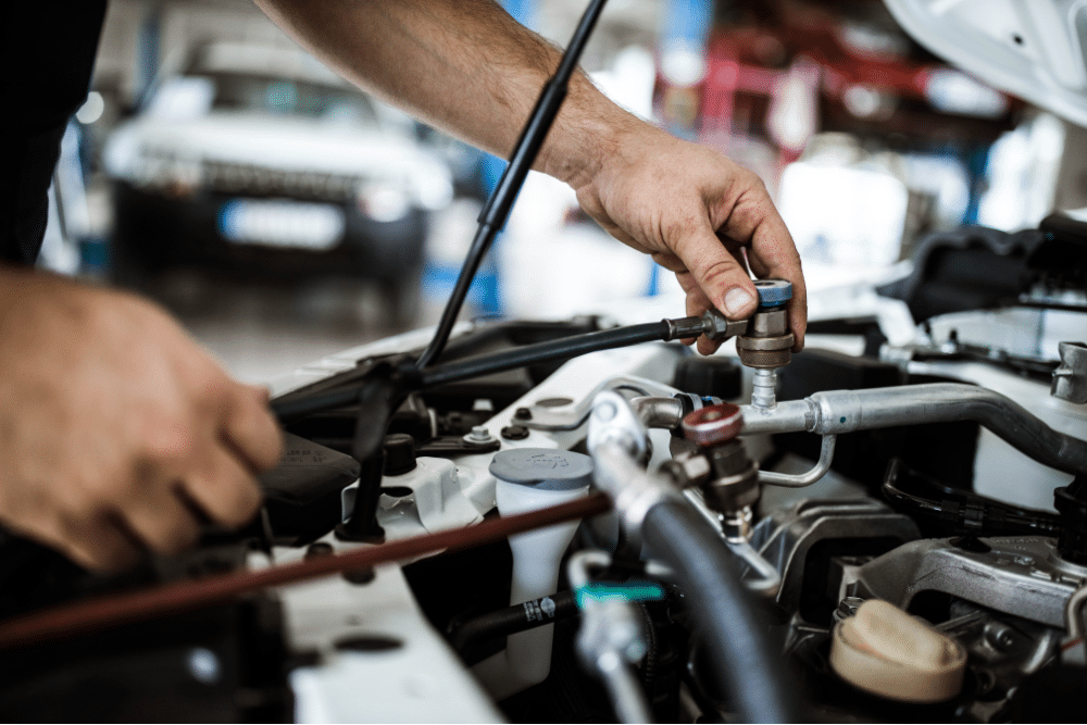 a/c recharge, auto repair in Milwaukee, WI at Next Level Auto Services. Technician attaching A/C service lines under the hood of a vehicle in a well-lit garage.