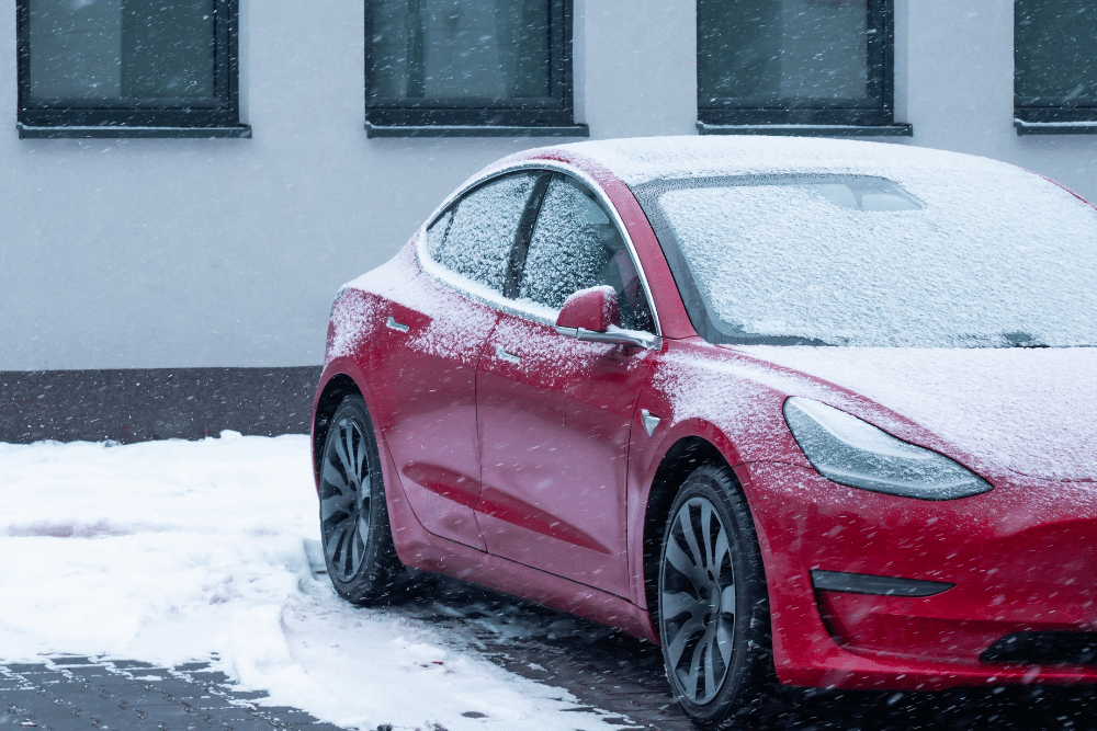 Tesla Maintenance in Milwaukee, WI by Next Level Auto Services. A red Tesla Model 3 parked outside in a snowstorm, with snow accumulating on the car and ground. The background shows a white building with dark windows, highlighting the winter conditions.